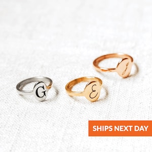 May include: Three gold rings with oval-shaped tops engraved with the letters G, E, and J. The rings are on a white fabric background.