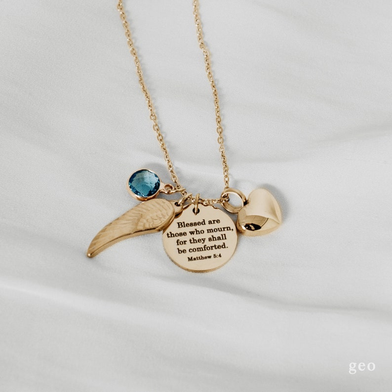 May include: Gold necklace with a charm that reads "Blessed are those who mourn, for they shall be comforted. Matthew 5:4". The necklace also features a small blue gemstone and a gold wing charm.