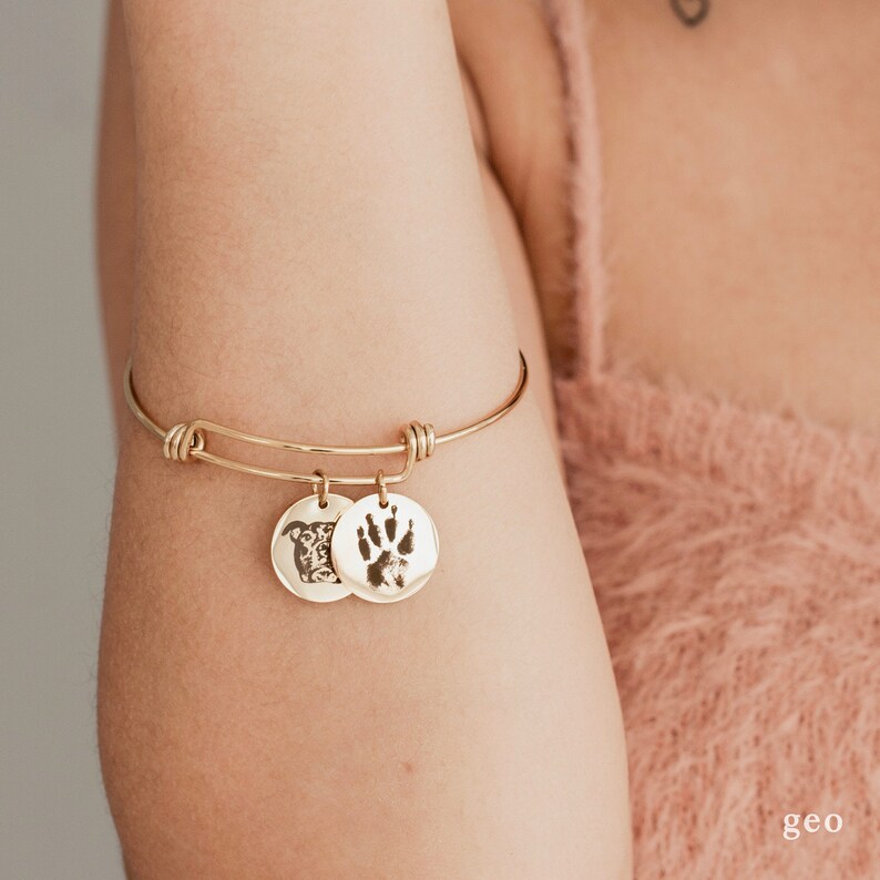 May include: A gold bangle bracelet with two charms. One charm is a round disc with a dog's face engraved on it. The other charm is a round disc with a paw print engraved on it.