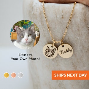 Op de afbeelding: Gouden ketting met twee ronde hangers. Een hanger heeft een kattengezicht gegraveerd, de andere hanger heeft de naam "Maddy" gegraveerd. De ketting is afgebeeld tegen een lichtbruine achtergrond.  Graveer je eigen foto!  Verzending de volgende dag.