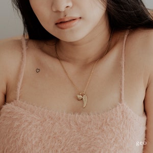 May include: A gold chain necklace with a small heart charm and a larger wing charm. The necklace is worn by a person with a small heart tattoo on their chest.
