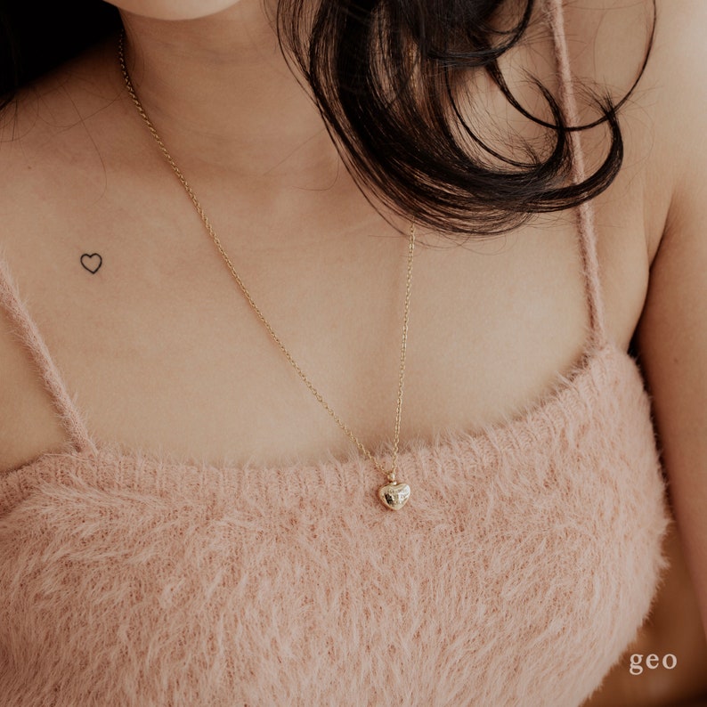 May include: A gold chain necklace with a small, heart-shaped pendant. The necklace is worn by a person with a small heart tattoo on their chest.