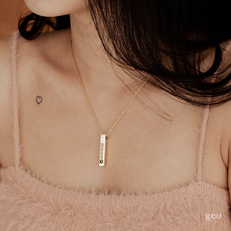 May include: A gold necklace with a vertical bar pendant engraved with the date "3.1.10.2.14.20" and paw prints.