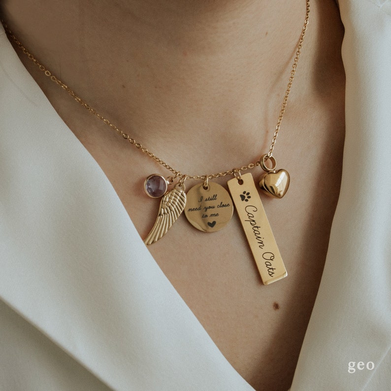 May include: Gold necklace with multiple charms, including a heart, a paw print, a wing, and a round charm with the inscription "I still need you close to me". The bar charm has the inscription "Captain Oats".