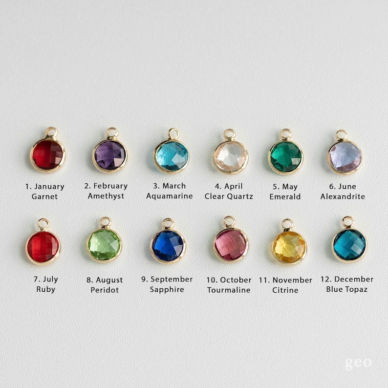 May include: Twelve gold charms with different colored gemstones, each representing a different month of the year. The charms are arranged in two rows of six. The first row includes garnet, amethyst, aquamarine, clear quartz, emerald, and alexandrite. The second row includes ruby, peridot, sapphire, tourmaline, citrine, and blue topaz.
