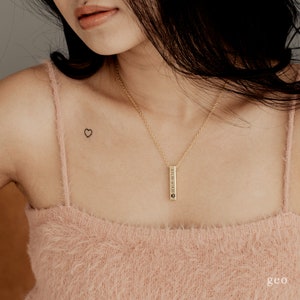 May include: A woman wearing a light pink fluffy tank top with a gold chain necklace and a small vertical bar pendant. The pendant has a paw print and the text "2019.01.14.2021" engraved on it. The woman has a small heart tattoo on her left shoulder.