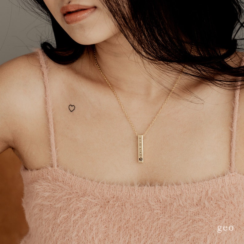 May include: A woman wearing a light pink fluffy tank top with a gold chain necklace and a small vertical bar pendant. The pendant has a paw print and the text "2019.01.14.2021" engraved on it. The woman has a small heart tattoo on her left shoulder.
