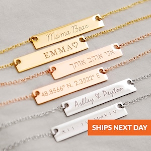 May include: A collection of personalized bar necklaces in gold, rose gold, and silver. Each necklace features a rectangular bar pendant with engraved text, including names, coordinates, and phrases like "Mama Bear" and "EMMA". The necklaces are displayed on a light gray surface.