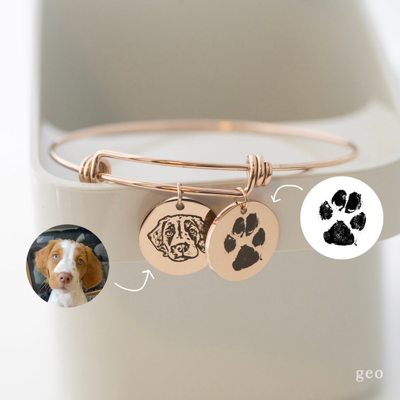 May include: A gold adjustable bangle bracelet with two charms. One charm features a black and white engraved image of a dog's face. The other charm features a black and white engraved paw print.