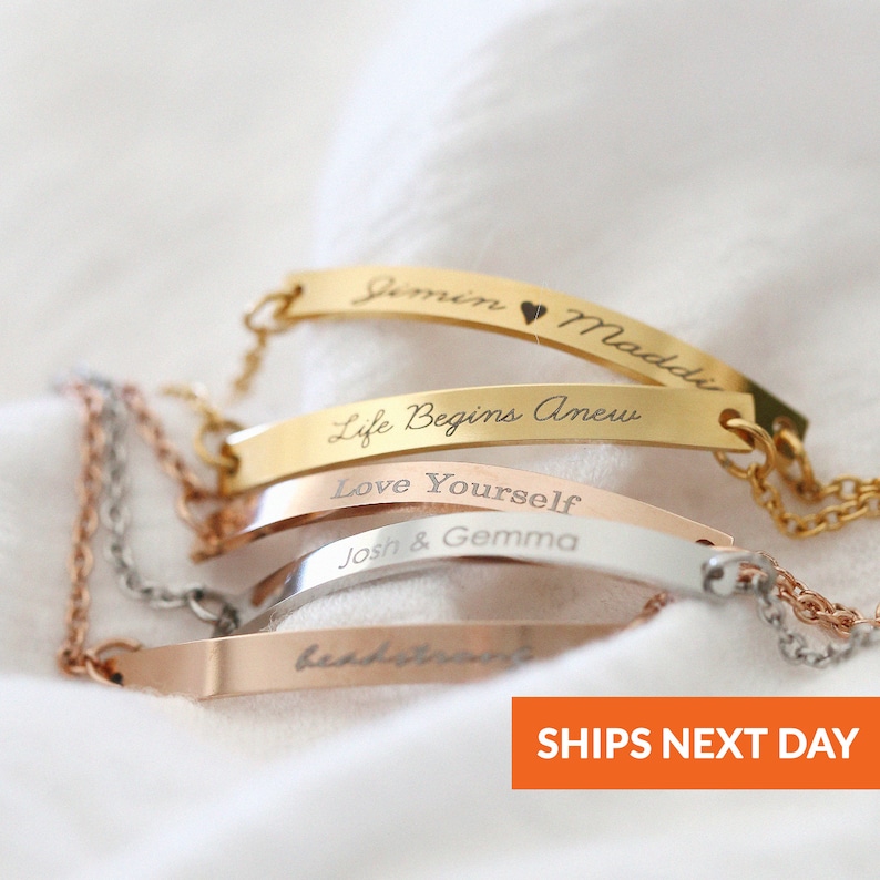 May include: Four gold, silver, and rose gold bracelets with engraved messages. The bracelets feature a delicate chain and a curved bar with engraved text. The text on the bracelets reads "Jimin Maddie", "Life Begins Anew", "Love Yourself", and "Josh & Gemma".