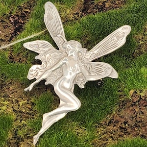 May include: A silver fairy figurine with outstretched wings and flowing hair, resting on a bed of green grass. The fairy is depicted in a graceful pose, with intricate details in the wings and body.
