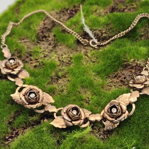 May include: A gold-toned necklace featuring rose-shaped pendants with small, sparkling center stones. The necklace has a delicate chain and is set against a backdrop of green moss. The rose pendants are detailed with leaves.