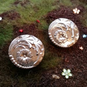 May include: Two silver-colored, round buttons with floral and leaf designs. The buttons have a detailed, embossed pattern featuring a central flower and surrounding foliage. The buttons are set against a backdrop of brown and green textured material with small decorative flowers.