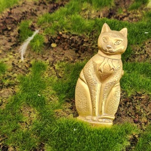 May include: A golden cat figurine with a detailed design, sitting upright. The cat has a scarf around its neck and is set against a backdrop of green grass and soil. The figurine is made of a shiny metal.