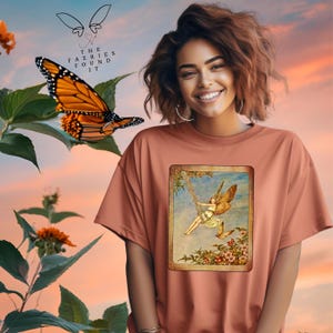 May include: A peach-colored t-shirt with a vintage illustration of a fairy with wings, sitting on a swing in a garden setting. The text "The Faeries Found It" is printed on the top of the shirt.
