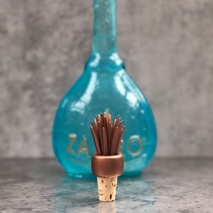Puede incluir: Una botella de vidrio turquesa con la palabra "ZARO" en letras doradas en el fondo. En primer plano, un tapón de botella con base de corcho, un anillo color cobre y un grupo de elementos marrones en forma de palito.