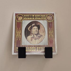 May include: A vinyl record album cover featuring Willie Nelson in a cowboy hat and a red and brown border. The album is displayed on a black wooden wall mount.