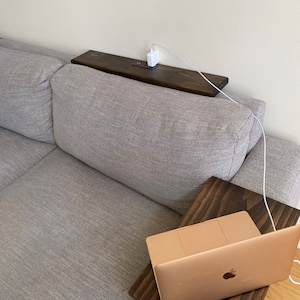 May include: A dark stained wooden sofa arm table with a built-in charging station. A white charging cable is plugged into the table and connected to a white power adapter. A laptop is on a matching wooden tray.