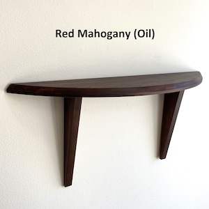 May include: A wall-mounted, semi-circular shelf made of red mahogany wood. The shelf has a curved front edge and is supported by two angled legs. The text "Red Mahogany (Oil)" is printed above the shelf.