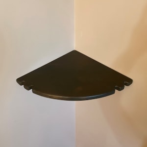May include: A dark brown, triangular corner shelf. The shelf has a curved front edge and decorative cutouts on the corners. It is mounted in the corner of a room where two walls meet.
