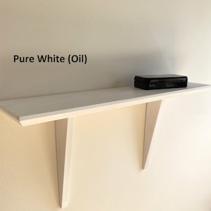 May include: A white floating shelf with two triangular brackets, mounted on a wall. The shelf is painted pure white and has a black box on top. The text "Pure White (Oil)" is visible on the wall.