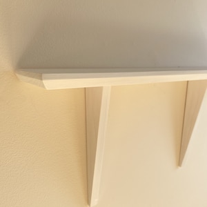May include: A light-colored wooden shelf mounted on a wall. The shelf has a simple, minimalist design with a flat top and angled supports. The shelf is approximately 30 inches long and is painted in a neutral color.