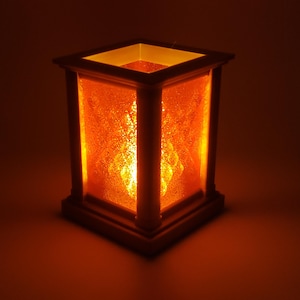 May include: A lit wooden lantern with a warm orange glow. The lantern has a square shape with textured glass panels and a decorative design in the center. The light source is inside, creating a cozy ambiance.