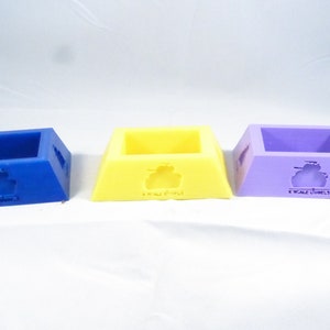 Puede incluir: Tres pequeños soportes de exhibición decorativos de forma cuadrada. Los soportes están hechos de plástico y son de color azul, amarillo y morado. Cada soporte tiene un pequeño diseño en relieve de un tanque en la parte delantera.
