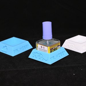 Pode incluir: Três suportes de plástico azul e branco com as palavras "MR. HOBBY" impressas neles. Um suporte contém uma garrafa transparente de cola com uma tampa roxa e as palavras "MR. HOBBY" e "CEMENTS SP" impressas no rótulo.