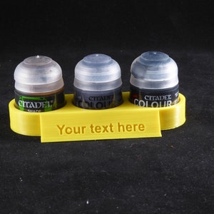 Pode incluir: Suporte para potes de tinta de plástico amarelo com três potes de tinta. Os potes de tinta são etiquetados como "Citadel Shade", "Citadel Colour" e "Citadel Colour". O suporte tem o texto "Your text here" impresso na frente.
