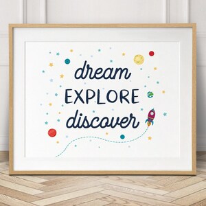 May include: A white framed print with a blue, yellow, and red rocket ship and stars. The text on the print reads "dream explore discover".