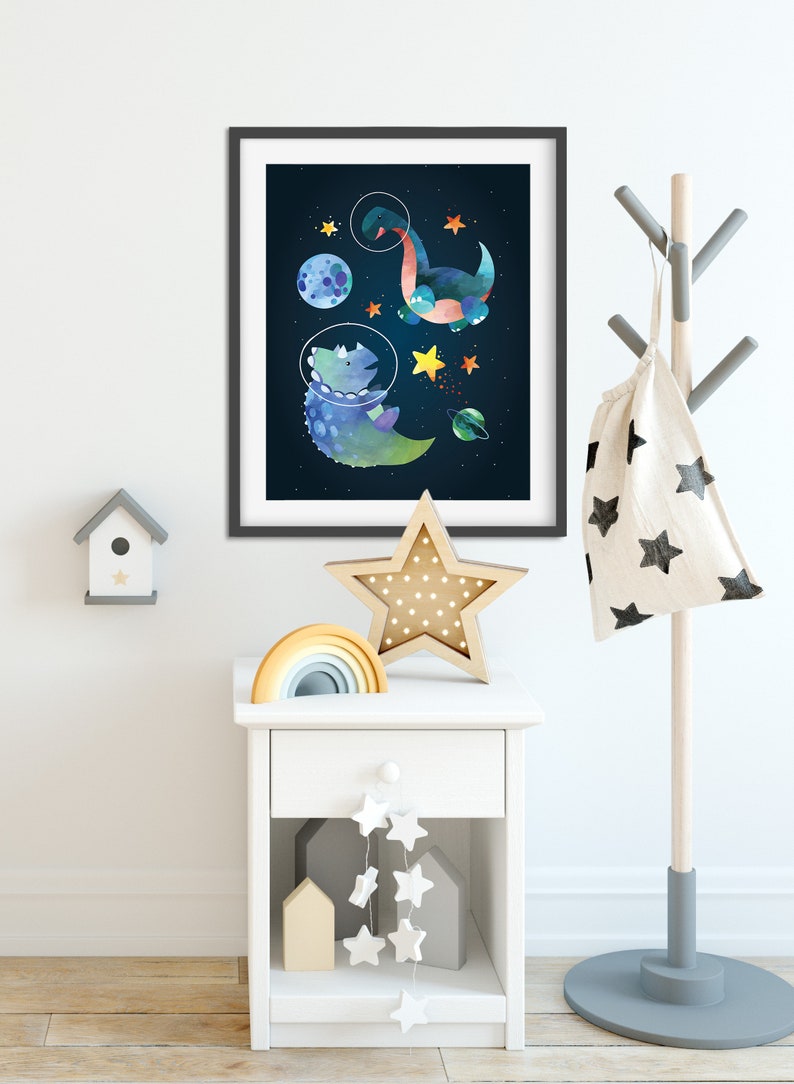 May include: A framed print of two dinosaurs in space suits floating in space with stars and planets. The print is hanging on a white wall above a white bedside table with a rainbow stacking toy, a star-shaped nightlight, and a string of white stars hanging from the bottom shelf.