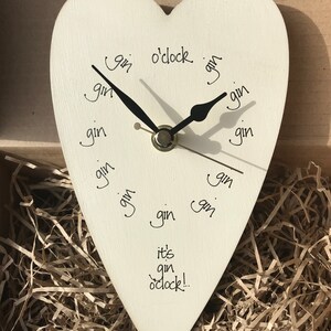 以下が含まれることがあります： ハート型のオフホワイトの時計。黒い針があり、「gin o'clock」と「it's gin o'clock!」の文字が書かれています。数字の代わりに「gin」の文字が使われています。時計は茶色の紙が入った箱に入っています。