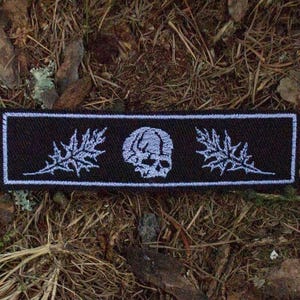 May include: A rectangular black patch with a white border, featuring a skull and leaf design. The patch is embroidered with white thread, and the design is centered on the patch. The patch is placed on a bed of brown leaves and pine needles.