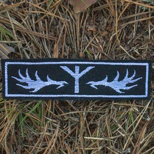 Op de afbeelding: Een rechthoekige zwarte patch met een wit geborduurd ontwerp. Het ontwerp heeft een centrale verticale lijn met een horizontale lijn aan de bovenkant, geflankeerd door gestileerde geweivormige vormen. De patch ligt op een achtergrond van bruine dennennaalden en groen gras.