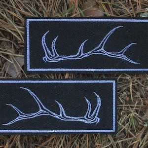 Puede incluir: Dos parches rectangulares negros con diseños de cuernos bordados en blanco. Los parches se muestran sobre un lecho de agujas de pino marrones y hierba seca. Los parches son de forma rectangular.