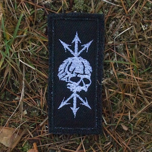 Pode incluir: Etiqueta bordada preta com um design de caveira e ossos cruzados brancos. A caveira é cercada por quatro flechas brancas apontando para fora.