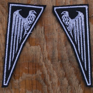 May include: Two black and white embroidered patches. Each patch is a stylized eagle with outstretched wings, set against a black background. The patches are triangular in shape with a white border. The patches are placed on a weathered wooden surface.