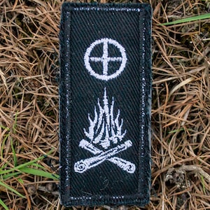 May include: A black rectangular patch with a white symbol of a circle with a cross on top, and a campfire design below. The patch is set against a backdrop of brown pine needles and green grass.