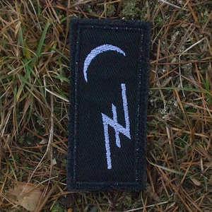 Op de afbeelding: Een zwart rechthoekig embleem met een wit maansikkel- en bliksemontwerp. De patch ligt op een bedje van bruine dennennaalden en gras.