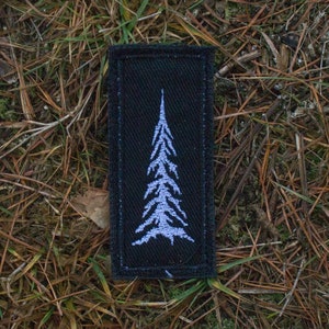 Puede incluir: Un parche de tela negro con una silueta bordada en blanco de un pino.