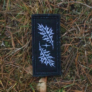 Puede incluir: Un parche rectangular negro con diseños de hojas bordadas en blanco. El parche presenta dos patrones de hojas en espejo, uno encima del otro, con una cruz en el centro. El parche está sobre un fondo de follaje marrón y verde.