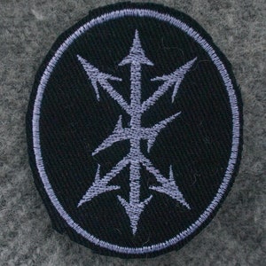 Puede incluir: Un parche bordado en blanco y negro con un diseño estilizado de flechas apuntando en diferentes direcciones. El parche es circular y tiene un borde blanco.