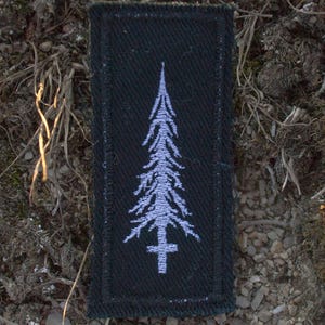 May include: A rectangular black patch featuring a white, stylized tree design with a cross at the base. The patch is set against a backdrop of dry earth, moss, and small stones, creating a natural contrast.