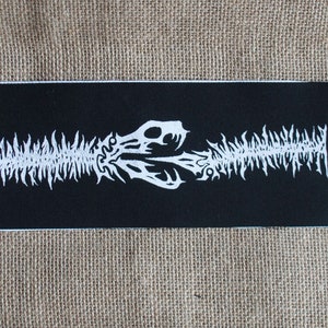 May include: Black patch with a white skeletal design of a creature with a long, segmented body and two heads. The design is symmetrical and has a jagged, spiky appearance.
