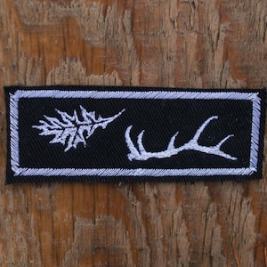Puede incluir: Un parche rectangular negro con un borde blanco. El parche presenta un diseño de hoja blanca a la izquierda y un diseño de cuerno blanco a la derecha. El parche está sobre un fondo de madera.