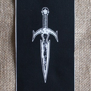 Puede incluir: Un parche de tela negro con un diseño de daga bordado en blanco. La daga tiene una punta puntiaguda y una guarda cruzada.
