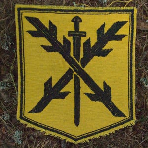 May include: A yellow patch with a black emblem featuring a sword and crossed stylized leaves. The patch has a black border and is set against a backdrop of forest floor with pine needles and twigs.