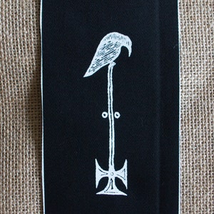 Könnte beinhalten: Schwarzer Stoff-Patch mit einem weißen gestickten Design eines Vogels, der auf einem Kreuz mit einem kreisförmigen Design an der Spitze des Kreuzes sitzt.