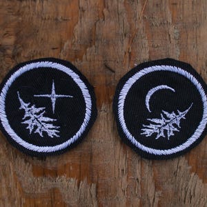 以下が含まれることがあります： 白のステッチが施された、2つの丸い黒色の刺繍パッチ。1つは十字架と木、もう1つは三日月と木が描かれています。風化した木製の表面に表示されています。